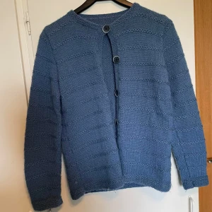 Blå stickad kofta med knappar - Säljer en mysig blå stickad kofta med dekorativa knappar framtill. Koftan har ett diskret randigt mönster och långa ärmar. Perfekt för kyliga dagar! Hemma stickad!