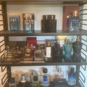 Parfymkollektion med olika märken - En samling av parfymer från olika märken, inklusive flaskor i blått, svart och genomskinligt glas. Märken som mancera och Riad syns tydligt. Flaskorna har olika former, från fyrkantiga till mer skulpturala designer. Perfekt för den som vill ha variation i sin doftgarderob. 