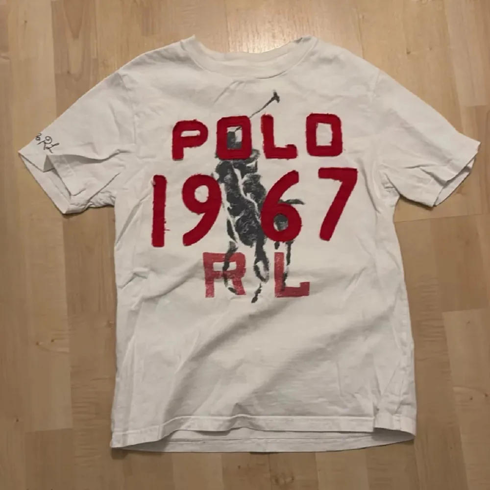 Säljer en vit t-shirt från Ralph Lauren med röd text och en stor svart häst- och ryttarmotiv på framsidan. T-shirten har korta ärmar och är i en klassisk passform. Perfekt för en avslappnad stil. Passar mer som xs . T-paidat.