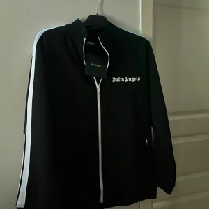 Svart track jacket från Palm Angels - Snygg svart track jacket från Palm Angels med vita detaljer och dragkedja. Jackan har en klassisk design med vita ränder längs ärmarna och Palm Angels-logga på bröstet. Perfekt för en stilren och sportig look. Helt oanvänd, lappen sitter kvar på den. Har resten av Dressen (dvs byxorna till zippen) i min profil om man skrollar