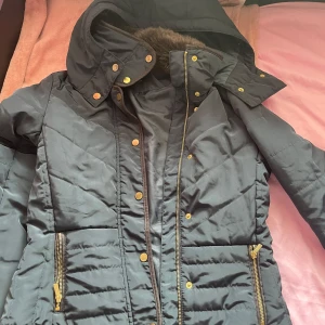 Svart quiltad jacka från H&M - Säljer en svart quiltad jacka från H&M med huva och dragkedja. Jackan har guldiga detaljer och en mysig fuskpäls på insidan av kragen. Perfekt för kyliga dagar!