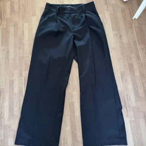 Svarta kostymbyxor från Gina Tricot - Snygga svarta kostymbyxor från Gina Tricot i storlek S. Byxorna har en klassisk design med mid waist och vida ben, perfekt för en stilren look. 👀 De är tillverkade i ett bekvämt material och har hällor för bälte.