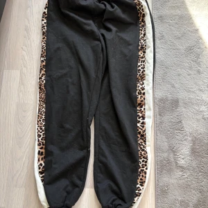 Svarta mjukisbyxor med leoparddetaljer - Snygga svarta mjukisbyxor med leopardmönstrade detaljer längs sidorna. Byxorna har en loose passform och är perfekta för en avslappnad stil. De är elastiska vid anklarna för extra komfort.