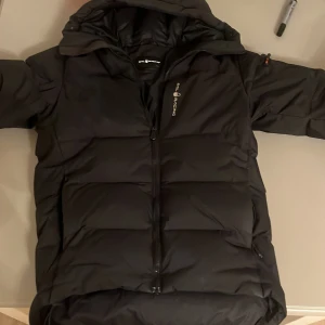 Svart dunjacka från Sail Racing - Säljer en svart dunjacka från Sail Racing. Jackan har en stilren design med dragkedja och huva. Perfekt för dig som gillar värme.
