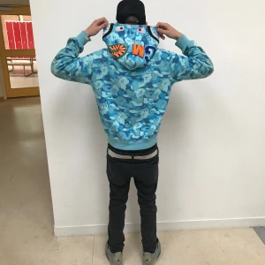 Blå camo hoodie med tryck - Säljer en cool blå camo hoodie med unikt tryck på huvan. Perfekt för dig som vill sticka ut med en streetwear-look. Hoodien har en dragkedja framtill och långa ärmar. Nytt sick storlek M passar S används it för att jag it gillar den 