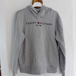 Grå hoodie från Tommy Hilfiger - Säljer en klassisk grå hoodie från Tommy Hilfiger med logotyp och årtal tryckt på framsidan. Tröjan har en bekväm passform med långa ärmar och en praktisk huva. Perfekt för en avslappnad stil. Sparsamt använd!