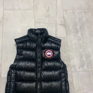 Canada Goose väst - Snygg svart dunväst med dragkedja och ett runt märke på bröstet. Perfekt för kyligare dagar och ger en stilren look. Västen har en hög krage för extra värme. Den är relativt ny köpt från Johnells för cirka 7000kr. Har även kvitto. Storlek Xs passar även S
