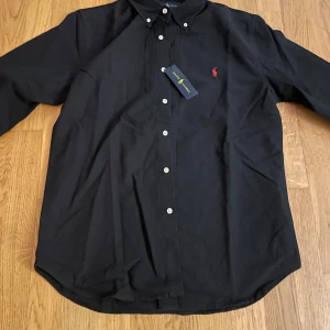 Svart skjorta från Ralph Lauren - Klassisk svart skjorta från Ralph Lauren med knappar framtill och röd broderad logga på bröstet. Skjortan har lång ärm och button-down krage, perfekt för en stilren look.