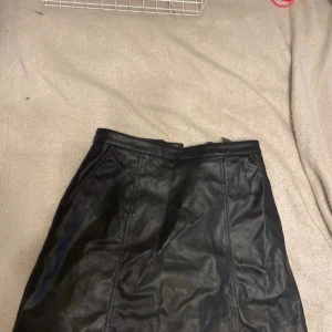 Svart skinnkjol från H&M Divided - Säljer en svart skinnkjol från H&M Divided i storlek 38. Kjolen har en rak passform, synliga sömmar och dragkedja bak. Knappt använd så i toppenbra skick, normal i storlek