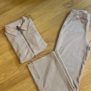 Beige ribbade mjukisbyxor med matchande ziptröja - Säljer ett par beige ribbade mjukisbyxor med resår i midjan och raka ben. Byxorna kommer med en matchande ziptröja med krage. Perfekt för en avslappnad och stilren look.