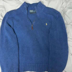 Polo Ralph Lauren Quarter Zip-up Blue - Mysig blå stickad tröja från Ralph Lauren med halv dragkedja och ribbade muddar. Klassisk gul logga broderad på bröstet och hög krage. Perfekt för kyliga dagar.