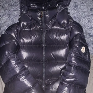 Mörkblå dunjacka från Moncler - Säljer en mörkblå dunjacka från Moncler med huva och dragkedja framtill. Jackan har ett klassiskt Moncler-märke på ärmen och quiltad design. Perfekt för kyliga dagar och håller dig varm med stil.