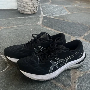 Svarta löparskor från Asics - Säljer ett par svarta Asics löparskor med vita detaljer och logga på sidorna. Skorna har snörning och en vit, tjock sula med svart mönster undertill. Perfekta för löpning eller träning.