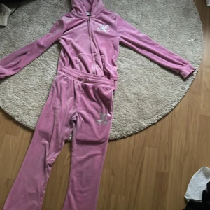 Rosa velour hoodie från Juicy Couture - Säljer en klassisk rosa velour hoodie från Juicy Couture med dragkedja och huva. Tröjan har långa ärmar och logga framtill samt texten 'Juicy' i glitter på ryggen. Perfekt för en avslappnad och trendig look. Och byxorna säljer jag också