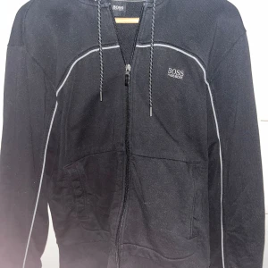Svart zip-hoodie från BOSS - Svart hoodie från BOSS med dragkedja framtill och vita kontrastsömmar. Justerbar huva med snören och två fickor på sidorna. Klassisk och stilren design med BOSS-logga på bröstet. Står L men sitter mer som M