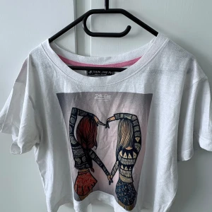 Vit croppad t-shirt med tryck - Vit croppad t-shirt med färgglatt tryck framtill som föreställer två tjejer som håller varandra i handen. T-shirten har korta ärmar och en avslappnad passform. Perfekt för en avslappnad och trendig stil.