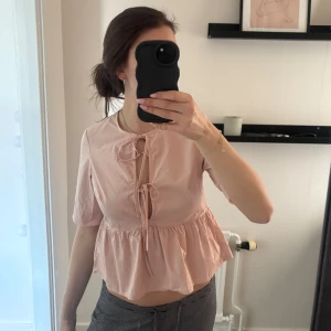 Ljusrosa blus med volang och knyt - Söt ljusrosa blus med volang nertill och knytning fram vid halsen. Blusen har korta ärmar och en luftig, lite lös passform. Perfekt för en somrig och feminin look.