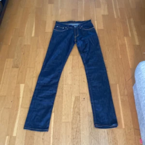 Mörkblå jeans från Nudie Jeans - Säljer ett par klassiska mörkblå jeans från Nudie Jeans med orangea sömmar och snygga broderade bakfickor. Byxorna har normal passform och raka ben. Perfekta för dig som gillar stilrena jeans med ikonisk design.