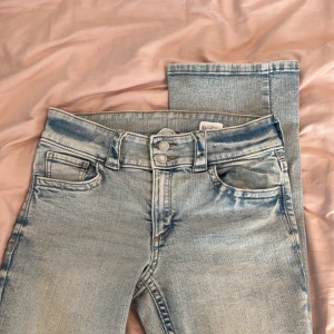 Ljusblå bootcut jeans från H&M - Säljer ett par ljusblå bootcut jeans från H&M med låg midja och justerbar midja. Jeansen har klassisk femficksdesign och knappar framtill. Perfekta för dig som gillar en avslappnad och tidlös stil.