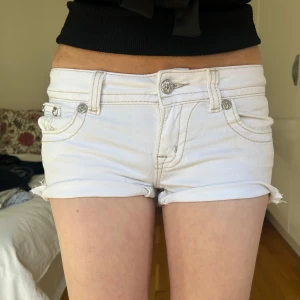 Vita jeansshorts från miss me - Säljer ett par vita, lågmidjade jeansshorts med rullade benslut och snygga silverfärgade nitar och broderier på bakfickorna. Shortsen har klassisk femficksdesign och dekorativa knappar. Perfekta för sommaren!