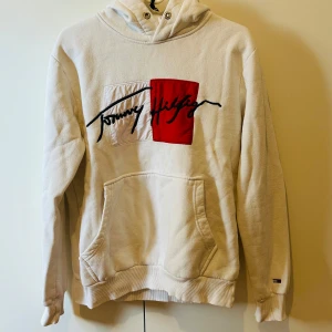 Vit hoodie från Tommy Hilfiger - Vit hoodie från Tommy Hilfiger med stor logga och röd detalj framtill. Klassisk känguruficka och huva med snörning. Passar egentligen storlek S 