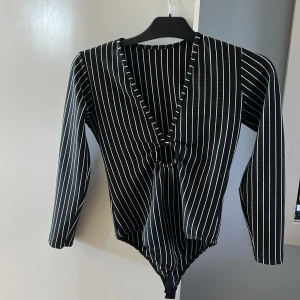 Svart och vit randig långärmad bodysuit - Säljer en svart bodysuit med vita vertikala ränder och djup v-ringning framtill. Bodyn har långa ärmar och knäppning i grenen. Perfekt för att styla med jeans eller kjol för en trendig look. Köpt i Brasilien och sedan dess bara legat i garderoben 