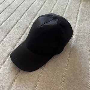 Svart keps från Gina Tricot - En stilren svart keps från Gina Tricot med klassisk böjd skärm och justerbar rem baktill. Kepsen är tillverkad i syntetmaterial och passar perfekt för en enkel och sportig look.