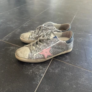Golden goose skor  - Säljer ett par glittriga sneakers från Golden Goose med silverfärgad paljett, rosa stjärna på sidan och grå snörning. Skorna har rund tå och vit sula med svarta detaljer vid hälen. Perfekta för dig som vill sticka ut!