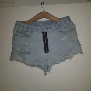 Ljusblå jeansshorts med nitar och dragkedja - Säljer ett par ljusblå jeansshorts med slitna detaljer, råa kanter och nitar längs midjan. Shortsen har en dekorativ dragkedja framtill med ring och är lågmidjade. Perfekta för sommaren!