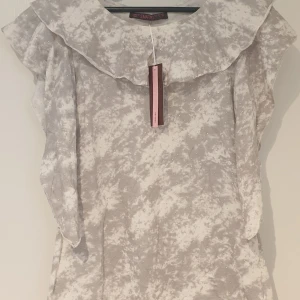 Linne med volang och pärlor från Semaforo - Supersött linne med volangkrage och pärldetaljer. Klänningen har ett diskret mönster perfekt för sommarens tillfällen. Materialet känns mjukt och lätt.