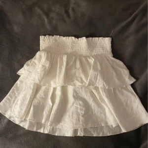 Vit volangkjol från SHEIN - Supersöt vit kjol från SHEIN med smock i midjan och dubbla volanger. Kjolen är kort och har en luftig känsla, perfekt för varma dagar. Enkel att matcha med olika toppar för en trendig look. Använd en gång, jättebra skick och normal i storlek