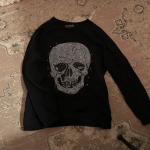 Svart tröja med döskalle från Saints & Mortals - Cool svart tröja från Saints & Mortals med en stor döskalle framtill i silvriga nitar och små guldstjärnor runtom. Perfekt statement-plagg för dig som gillar att sticka ut.