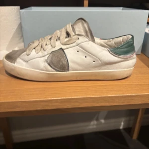 Vita sneakers från Philippe Model Junior - Säljer ett par vita sneakers från Philippe Model Junior med gråa och beiga detaljer. Skorna har snörning och en platt sula i gummi. Klassisk och stilren design med rund tå och diskreta färginslag. Skriv om ni har fler frågor eller om ni vill ha fler bilder. Lägg bud!