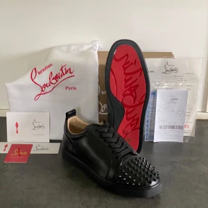 Christian Louboutin svarta läder - Säljer ett par svarta sneakers från Christian Louboutin med ikonisk röd sula och svarta nitar på tån. Skorna har snörning och är tillverkade i skinn. Perfekta för dig som vill sticka ut med en lyxig och edgy stil.