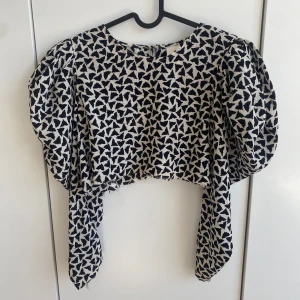 Svartvit mönstrad blus från H&M - Säljer en svartvit blus från H&M med grafiskt mönster och puffiga långärmar. Blusen har croppad passform och långa band framtill som kan knytas. Har klippt av den från en klänning så om man vill är det bara att sy upp där nere vid magen om man vill. Perfekt för dig som vill sticka ut med en unik topp!