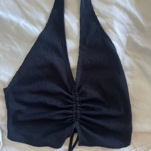 Svart ribbad halterneck topp - Säljer en svart ribbad halterneck topp med djup v-ringning och knytning framtill. Toppen har croppad passform och är ärmlös, perfekt för varma dagar eller en snygg layering-look. Endast använd 1 gång 