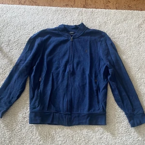Blå zip-jacka från Polo Ralph Lauren - Säljer en stilren blå jacka från Polo Ralph Lauren med dragkedja framtill och diskret broderad logga på bröstet. Jackan har lång ärm, ribbade muddar och är tillverkad i mjuk bomull. Perfekt för en avslappnad look.
