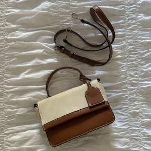 Brun och beige handväska med axelrem - Säljer en snygg handväska i brunt skinn och beige tyg. Väskan har både handtag och avtagbar axelrem, samt en dekorativ tag. Perfekt storlek för det viktigaste och stilren design.