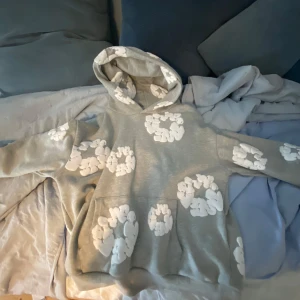 Grå hoodie med vita blommor från Denim Tears - Säljer en grå hoodie från Denim Tears med stora vita blommor tryckta över hela tröjan. Tröjan har huva och en stor magficka. Perfekt för dig som gillar streetwear och vill sticka ut lite extra.