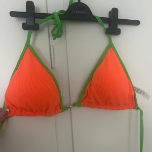 Orange och grön bikiniöverdel - Trendig bikiniöverdel i trekantsmodell med stark orange färg och gröna kanter. Knytband i nacken och ryggen för justerbar passform. Aldrig använd.