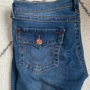 Blå jeansbyxor från True Religion - Säljer ett par jättesnygga blå jeans från True Religion med dekorativa bakfickor och kopparfärgade knappar💕💕Byxorna har låg midja och bootcut 🌸🌸 byxorna är använda fåtal gånger då de inte passar mig💕💕