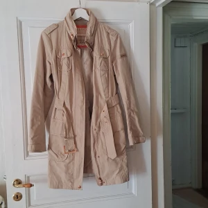 Beige trenchcoat med bälte - Snygg beige trenchcoat med klassisk krage och bälte i midjan. Jackan har flera fickor framtill, knappar och detaljer i samma färg. Perfekt för övergångssäsonger och ger en stilren look.