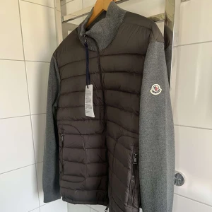 Grå dunväst med ärmar från Moncler - Snygg grå dunväst från Moncler med stickade ärmar och hög krage. Västen har två dragkedjefickor framtill och Moncler-logga på vänster ärm. Framsidan är quiltad och stängs med dragkedja. Perfekt för lager-på-lage -stil. Jackan är nästan helt o använd och kommer med kvitto. 