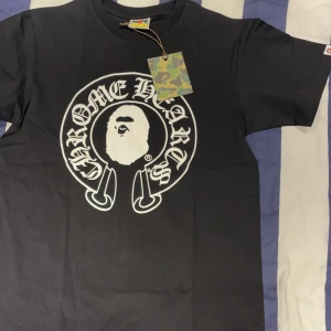 Svart t-shirt från Chrome Hearts x A Bathing Ape - Säljer en svart t-shirt från det coola samarbetet mellan Chrome Hearts och A Bathing Ape. T-shirten har ett stort tryck med 'Chrome Hearts' och 'Baby Milo' i vitt på både fram- och baksidan. Den är kortärmad och perfekt för en avslappnad stil perfekt till sommar 