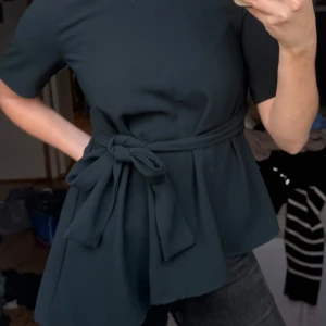 Mörkgrön blus med knytdetalj - Säljer en mörkgrön blus med korta ärmar och avtagbart knytband i midjan. Blusen har en lätt utsvängd passform och är tillverkad i ett mjukt tyg som ger ett stilrent intryck.