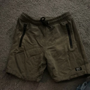 Olivgröna shorts med dragkedjor från KappAhl - Säljer ett par olivgröna shorts från KappAhl med svarta dragkedjor på fickorna och snörning i midjan. 