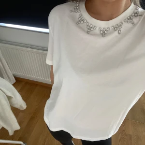 Vit t-shirt med strassdetaljer vid halsen - Säljer en vit t-shirt med korta ärmar och snygga strassdetaljer runt halsringningen. 