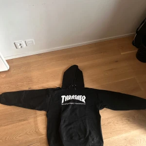 Svart Thrasher hoodie - Säljer en svart hoodie från Thrasher med vit logga på bröstet. Klassisk modell med huva och magficka, perfekt för skate eller streetwear. Tjockt och mjukt material som håller dig varm.