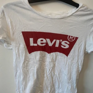 Vit t-shirt med röd Levi's-logga - Vit t-shirt från Levi's med den klassiska röda loggan tryckt framtill. T-shirten har rund halsringning och korta ärmar. Perfekt för en avslappnad och stilren look.Frakt tillkommer