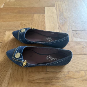Coola jeansballerinaskor  - Snygga ballerinaskor i blått jeansmaterial från Tiamo med spetsig tå. Skorna har dekorativa guldknappar och dragkedjedetaljer framtill samt kontrastsömmar. Perfekta för dig som vill ha en unik touch på outfiten. Står att skorna är i stlk 37 men skulle säga att de är något små i storlek!